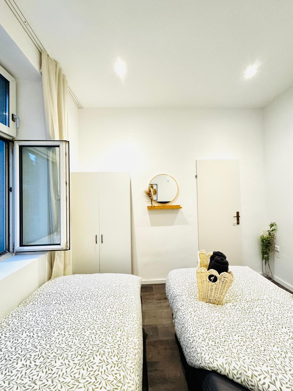 A hotel room with two beds, a window with curtains, a mirror on the wall, and a small shelf with a plant.