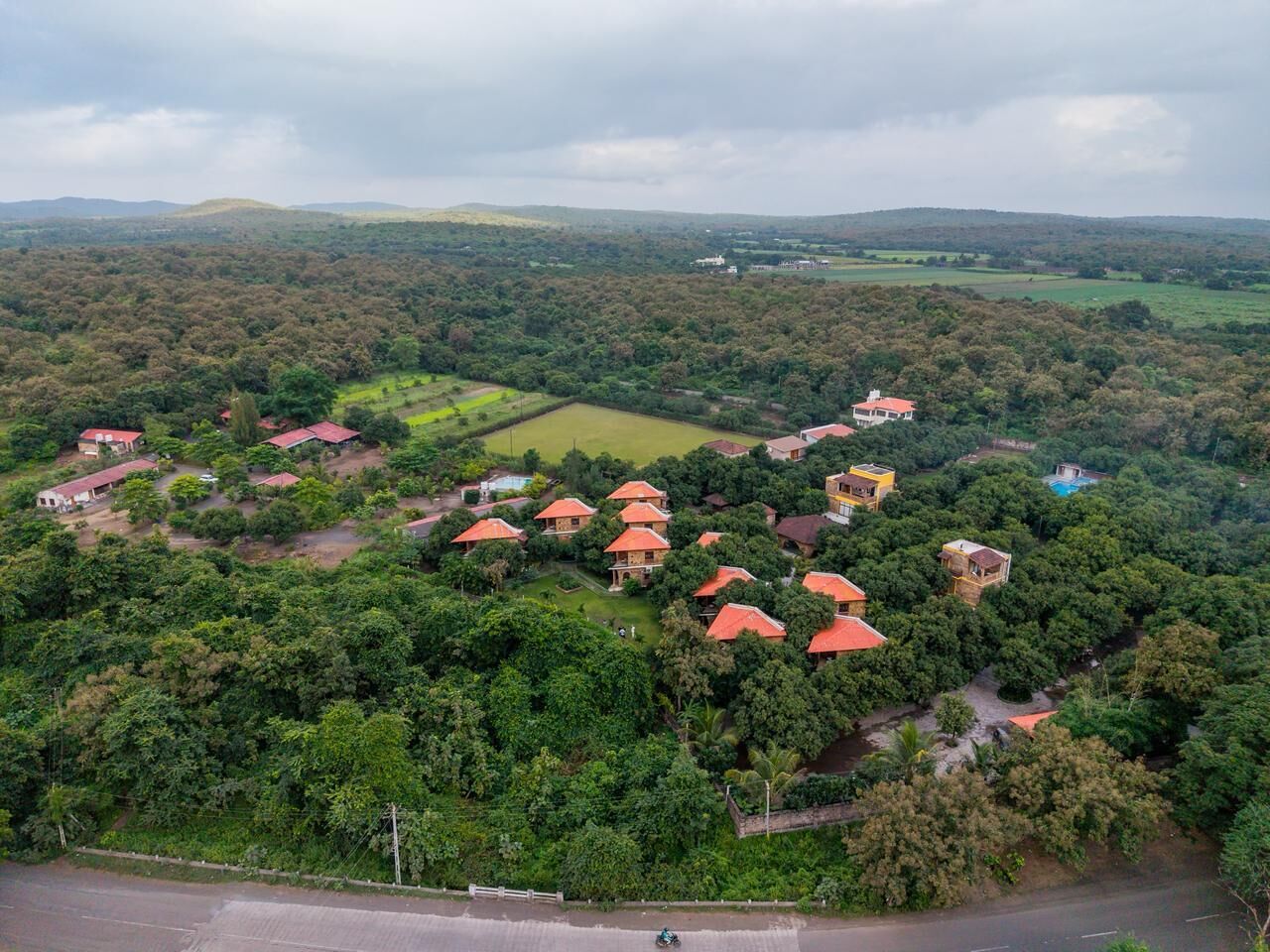 An aerial view of a residential area with multiple houses, some with red roofs, surrounded by dense greenery and fields.