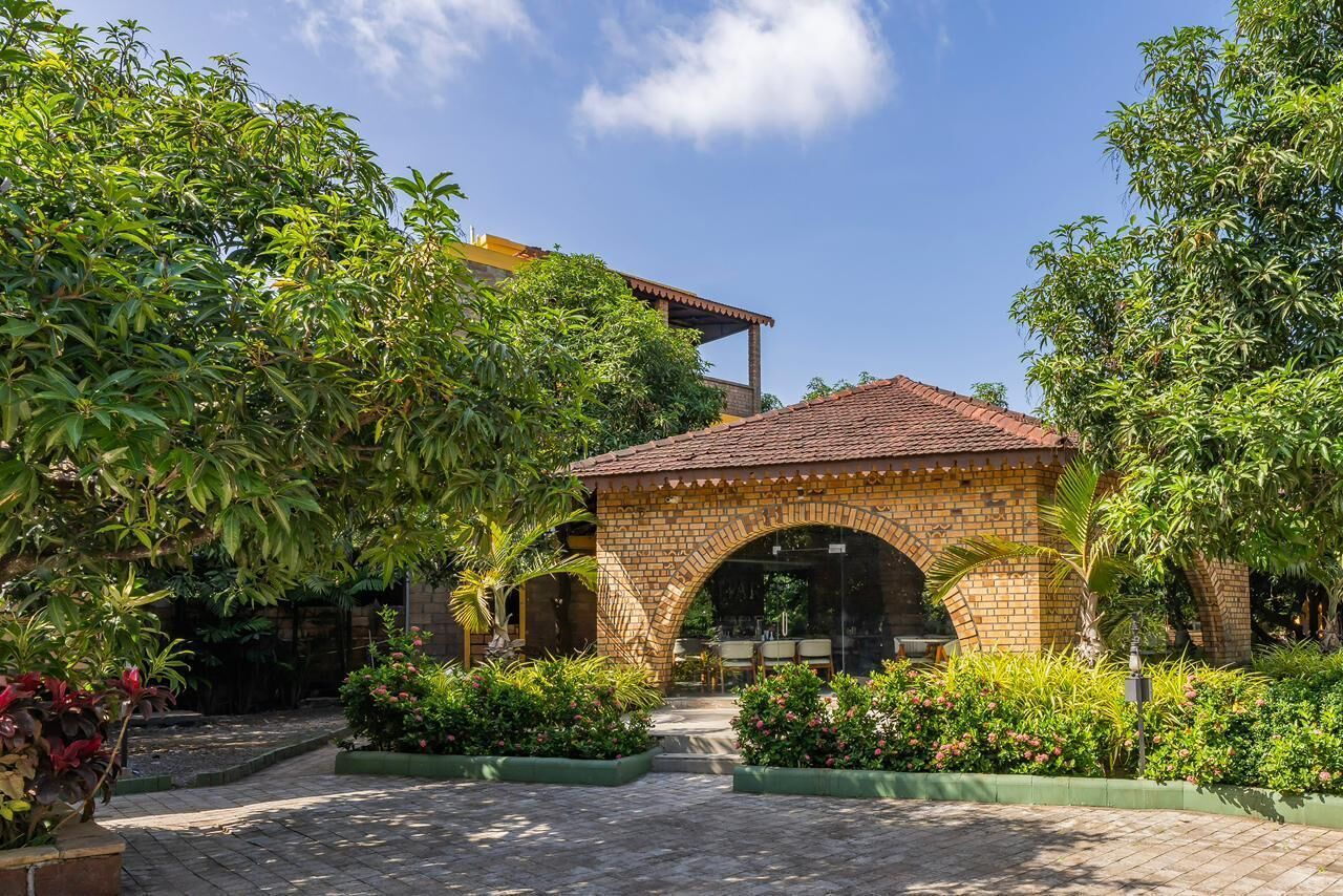 Un patio con un arco de ladrillo que conduce a una zona de estar cubierta, rodeado de abundante vegetación y un camino adoquinado.