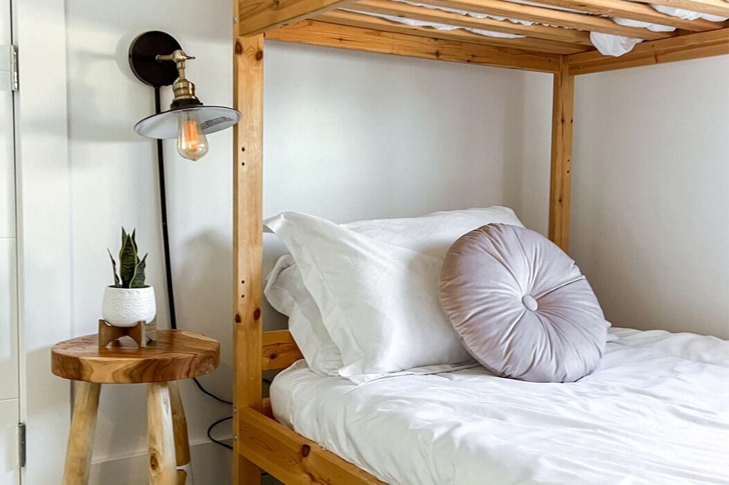Una cama individual con ropa blanca de cama y un cojín rosa, un taburete de madera pequeño con una planta en maceta, y una lámpara de pared.