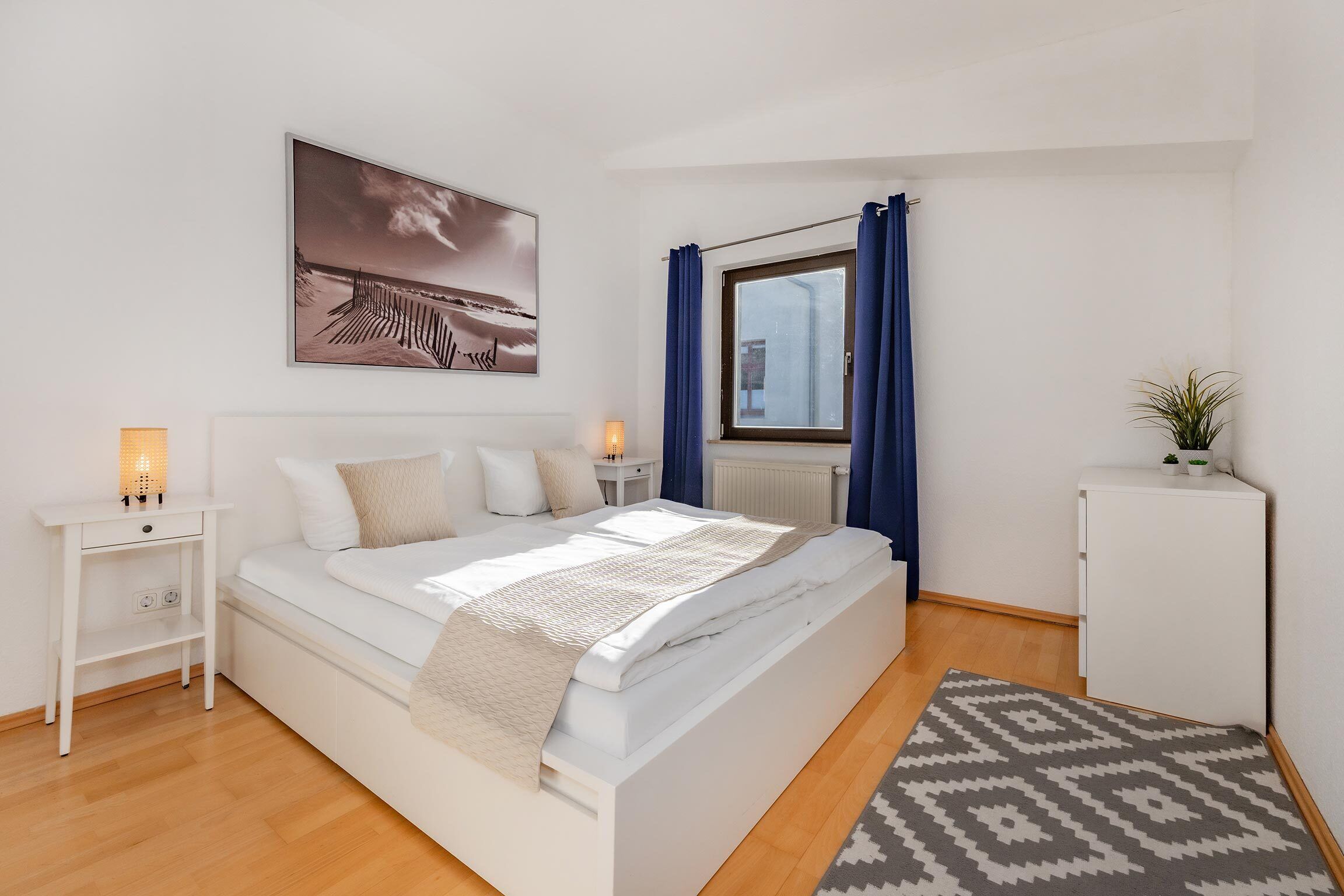 A neatly arranged bedroom with a bed, bedside tables, a radiator, a window with curtains, and a small cabinet.