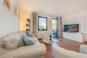 Un soggiorno moderno con un divano beige, una poltrona bianca, un piccolo tavolino con una bottiglia e dei bicchieri, una TV a schermo piatto su un mobile bianco e un'ampia finestra con vista all'esterno.