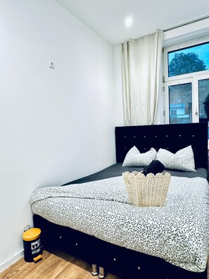 Ein Schlafzimmer mit einem Bett, einem kleinen Hocker und einem Fenster mit Vorhängen.