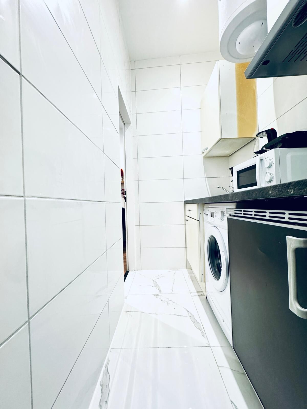 Una cocina pequeña con azulejos blancos, lavadora, microondas y pileta.