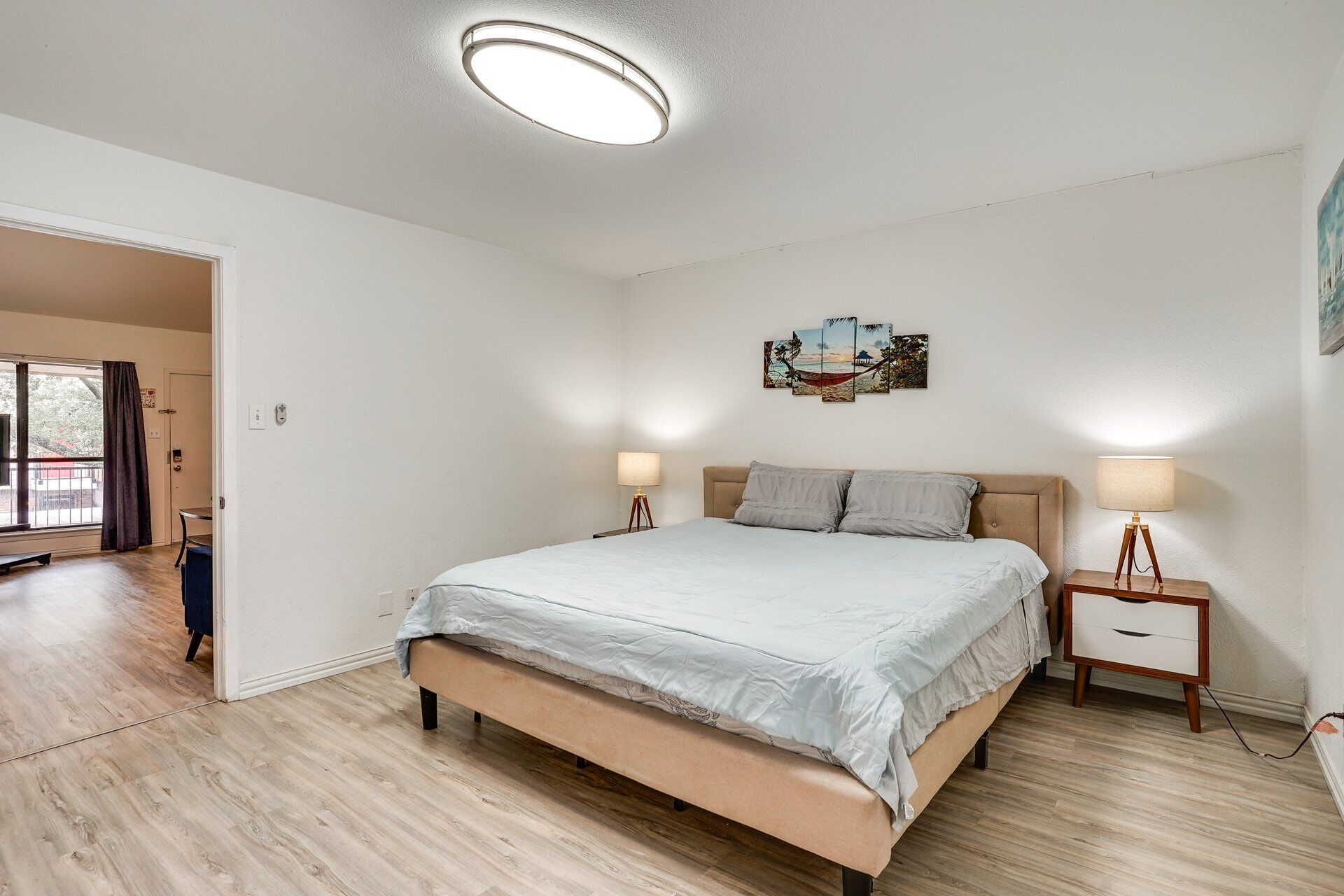 Un dormitorio con cama, mesitas de noche, una lámpara en el techo y una puerta que da a otra habitación.