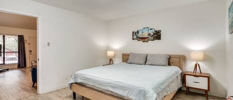 Un dormitorio con cama, mesitas de noche, una lámpara en el techo y una puerta que da a otra habitación.