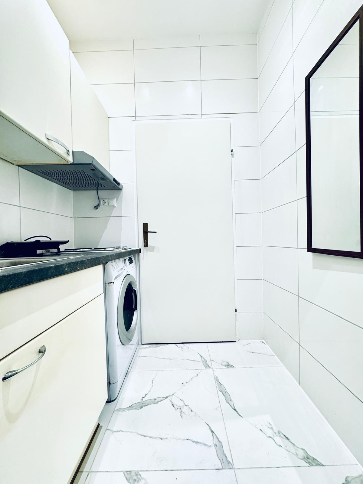 A compact kitchen with a built-in washing machine, white tiled walls, and a marble floor.