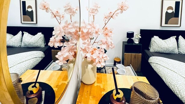 Een moderne slaapkamer met een grote spiegel, een vaas met roze bloemen, een houten nachtkastje en een bed met beddengoed met een patroon.