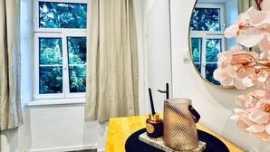 Een kamer met een houten tafel, een ronde spiegel, een raam met gordijnen en uitzicht op groen.