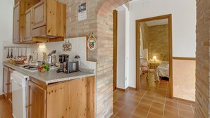 Cuina amb armaris de fusta, taulell de granat i paret de maó. Hi ha una zona de menjador amb taula i cadires, i un dormitori amb llit i una meseta de nit.