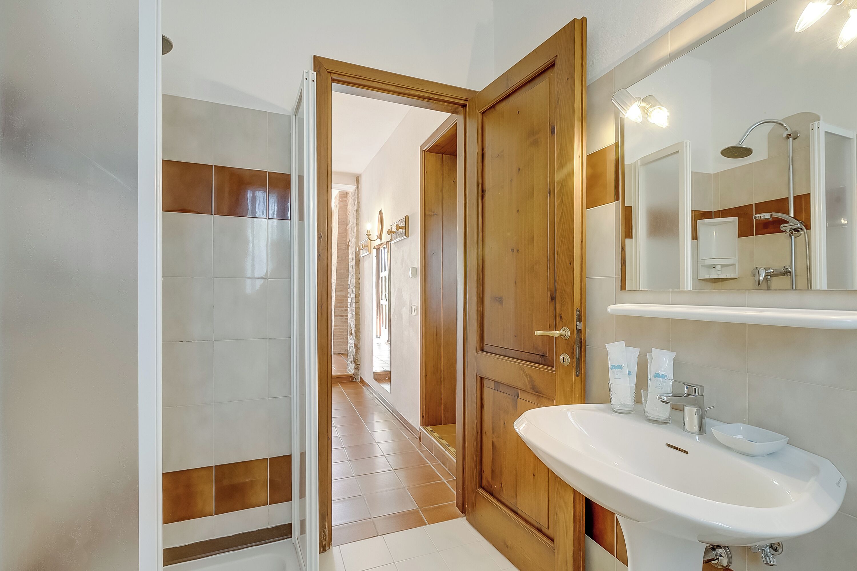 Une salle de bain moderne avec une porte en bois, des carreaux blancs, un grand miroir et un lavabo avec des articles de toilette.