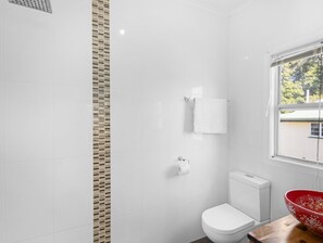 A modern bathroom with white tiles, a toilet, a red bowl, and a window with a view of trees. - Spacious Home Near Urangan Beachfront (Hervey Bay)