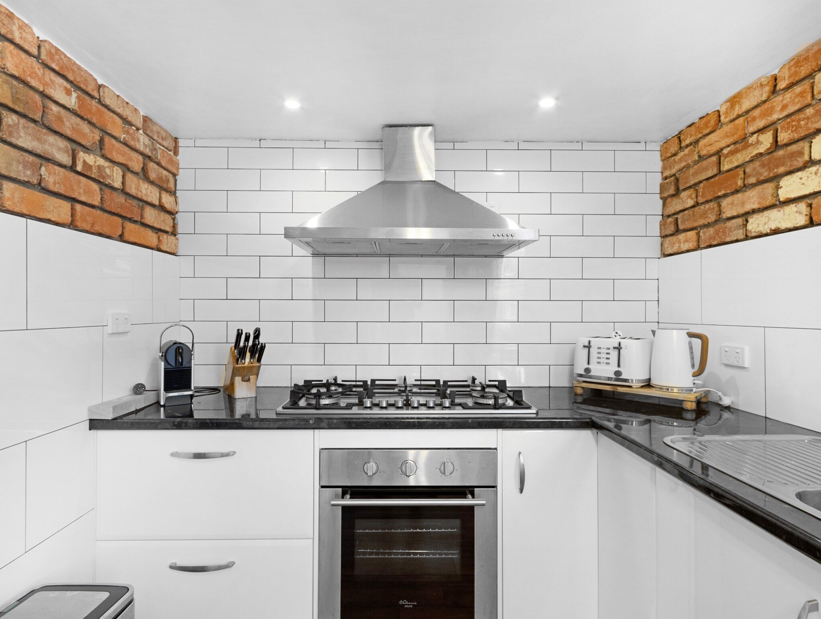Una cocina moderna con armarios blancos, extractor de acero inoxidable y pared decorativa de ladrillo.