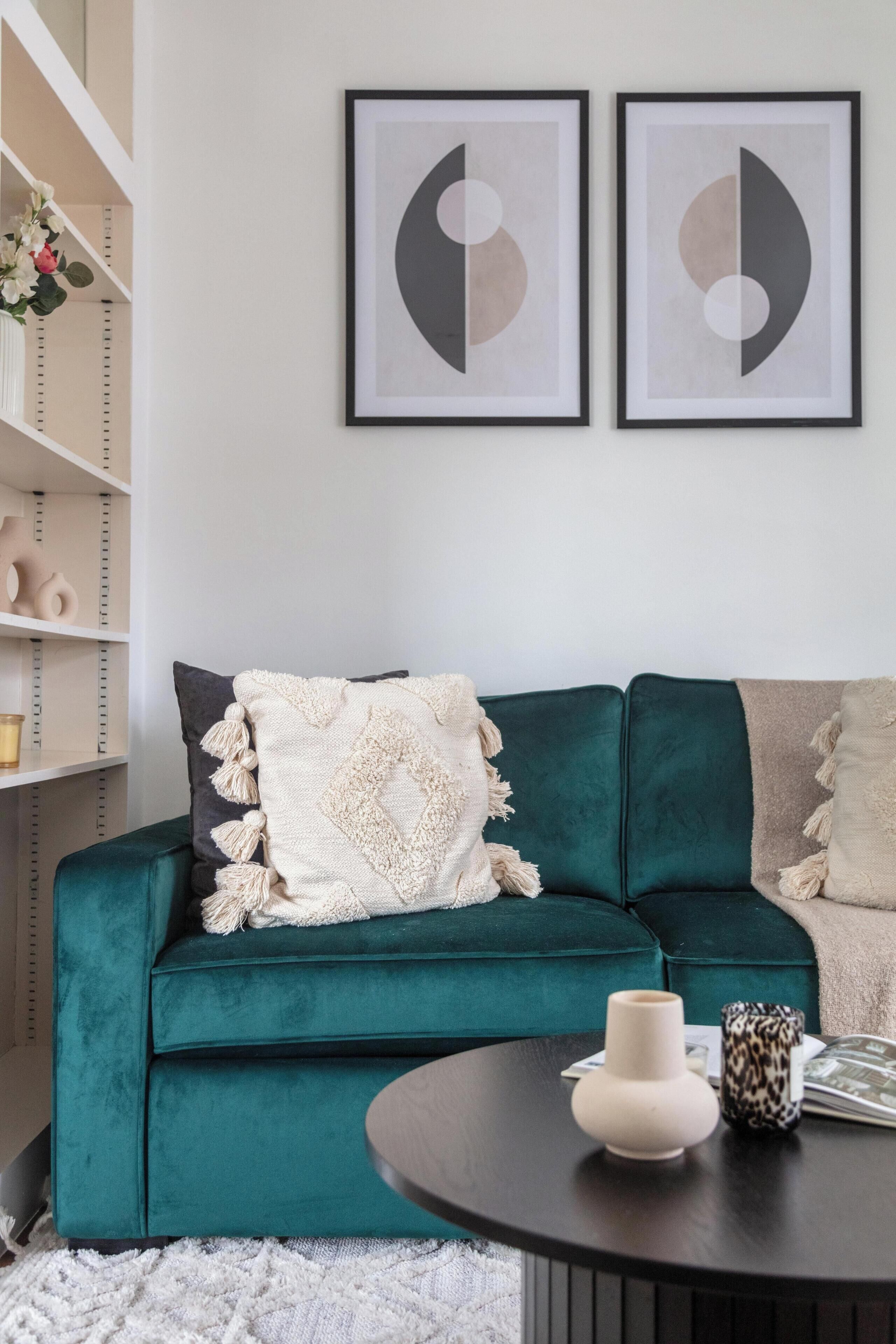 A teal sofa with a white throw blanket, a black coffee table with a white vase and a patterned mug, and a bookshelf with decorative items.
