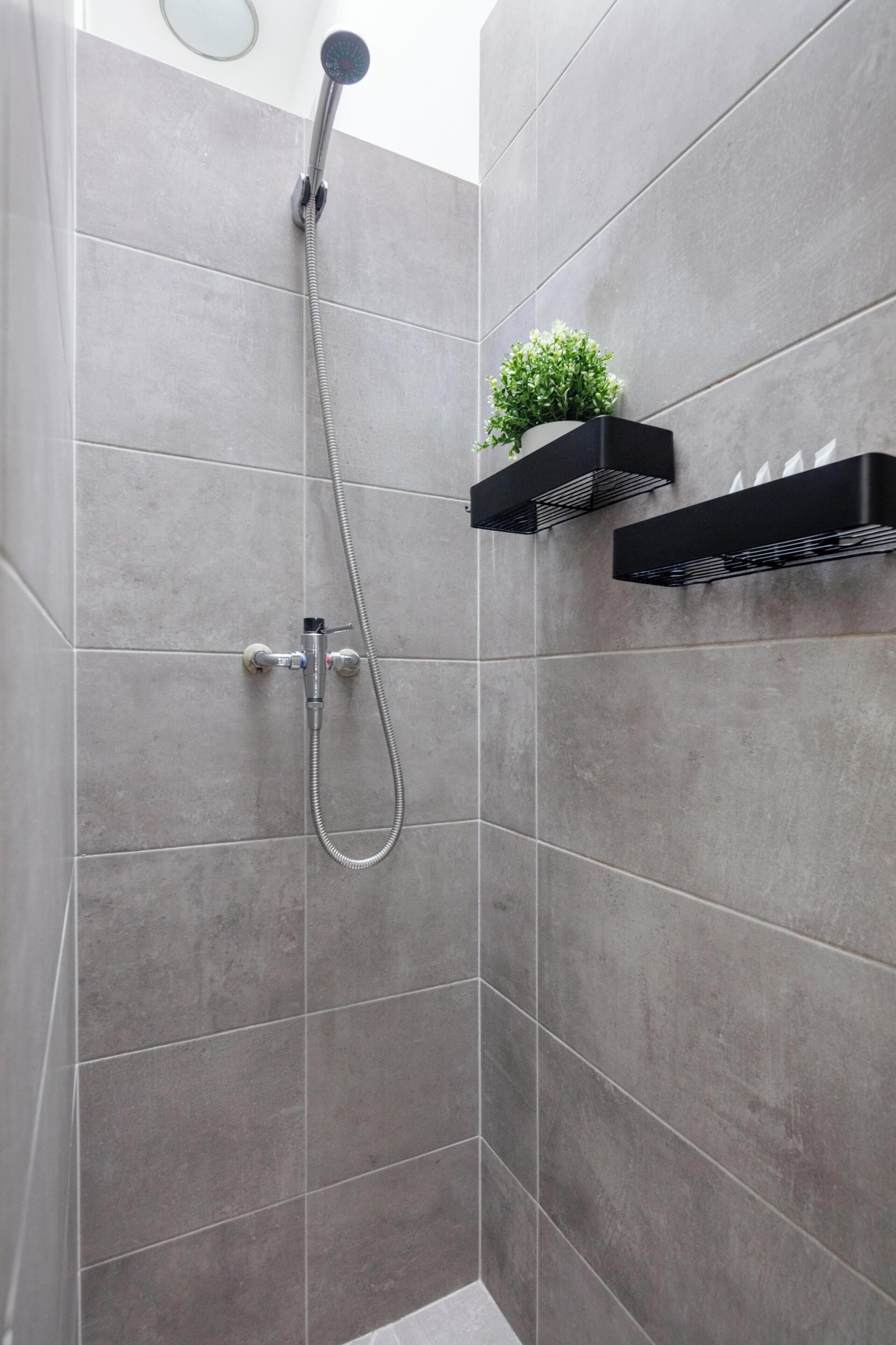 A shower area with grey tiles, a handheld showerhead, and a small plant on a black shelf.