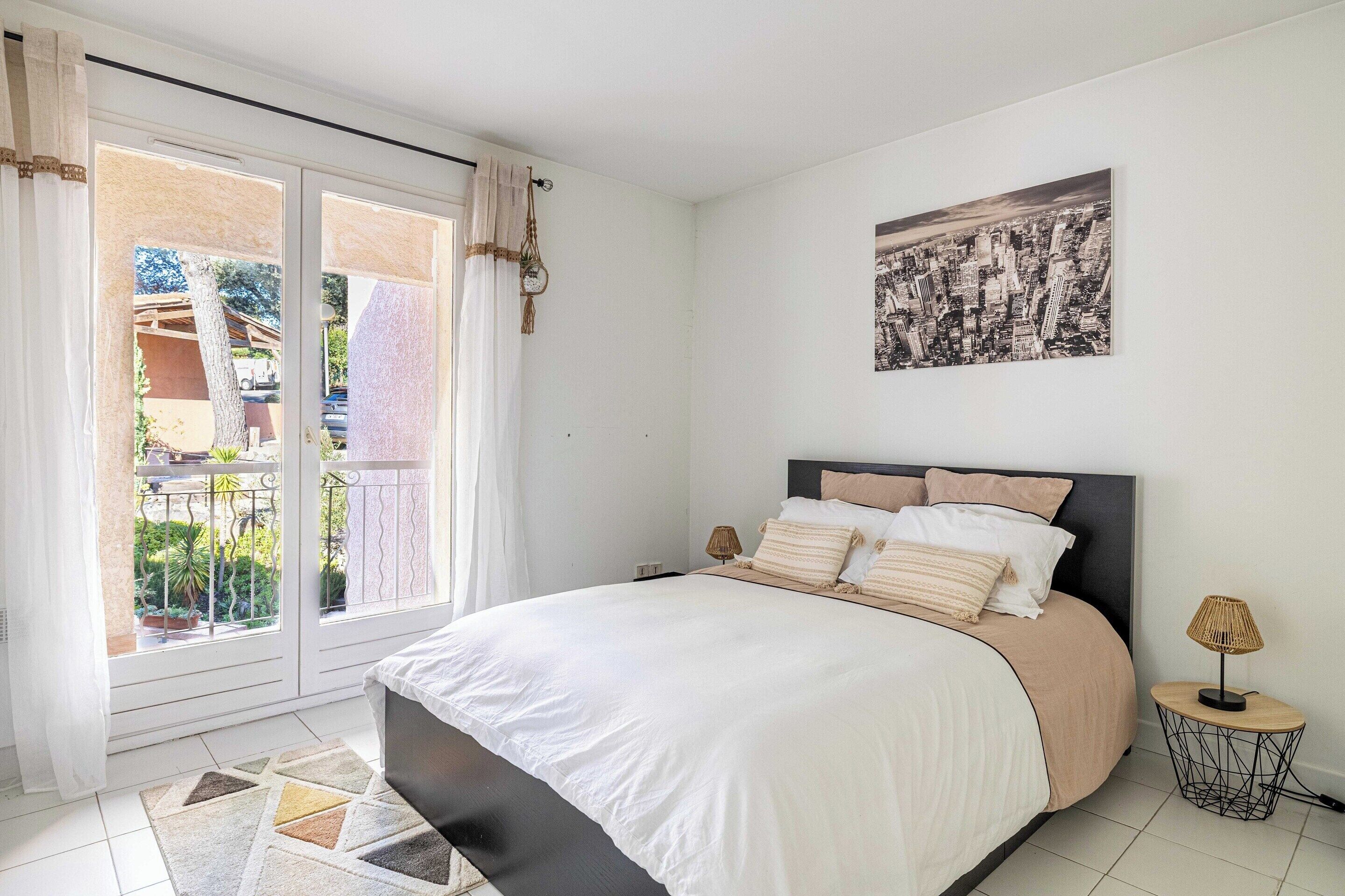 A bedroom with a bed, a nightstand, a lamp, a cityscape painting, and a view of a balcony with plants and a car.