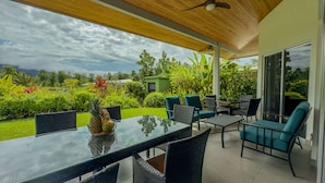 Un patio exterior con una mesa de vidrio, sillas de mimbre y vistas a un jardín exuberante y montañas.
