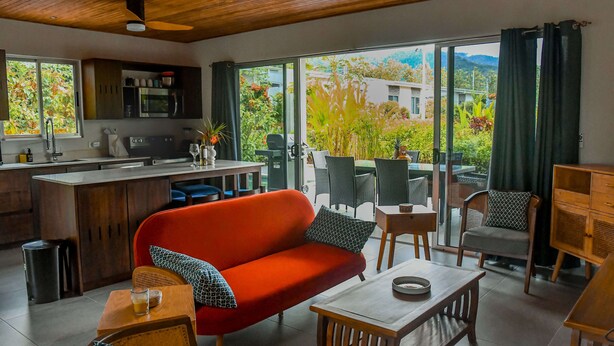 Una cocina moderna con comedor, un sofá rojo y vistas a un jardín exuberante a través de amplios ventanales correderas.