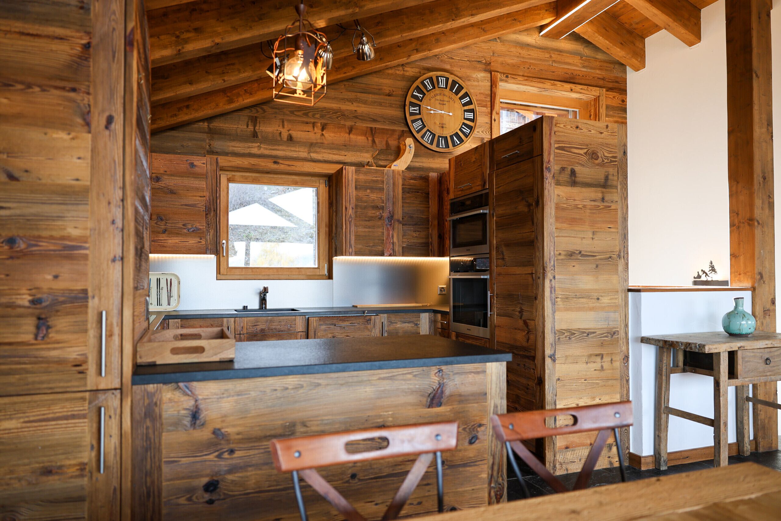 A rustic wooden interior with a wooden clock, a window, and a kitchen area with a sink and cabinets.