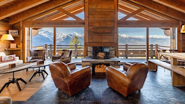 Een knus interieur van een lodge met een stenen openhaard, lederen fauteuils en een groot raam met uitzicht op besneeuwde bergen.