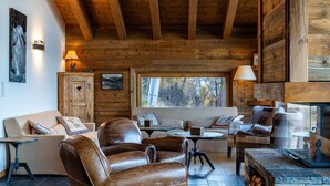 Een gezellig interieur van een blokhut met houten balken, leren banken, een stenen openhaard en een groot raam met uitzicht op het bos.