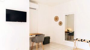 A modern, well-lit room with a flat-screen TV, a wooden desk, a grey chair, a small trash bin, and a wooden drying rack. - Florida Ascona Rooms (Ascona)