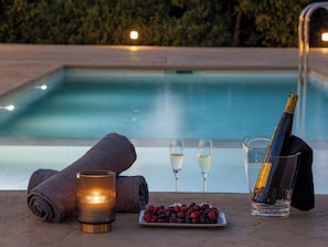 Escena junto a la piscina con una botella de champán, dos copas, una vela y un plato de uvas.