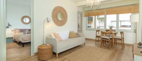 A modern living room with a sofa, dining table, and chairs. There is a chandelier, a mirror, and a window with blinds.