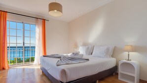 A bedroom with a large bed, a nightstand, a lamp, and a view of the sea through a window. - Casa Laranja by HR Madeira (Tabua)