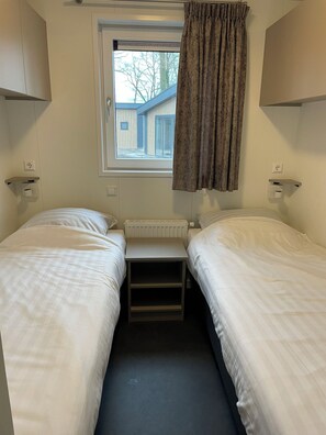 A small, single-room with two beds, a window with a view of the outside, and a radiator under the window. - WoodCottage for 6 people in the vacation park on the edge of Springendal in Ootmarsum (Oud Ootmarsum)