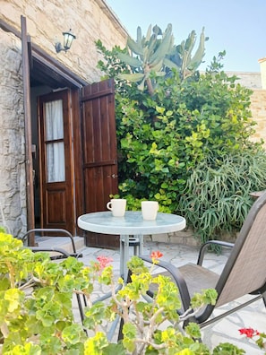 Zona exterior amb una taula rodona i cadires, una porta de fusta i diverses plantes.