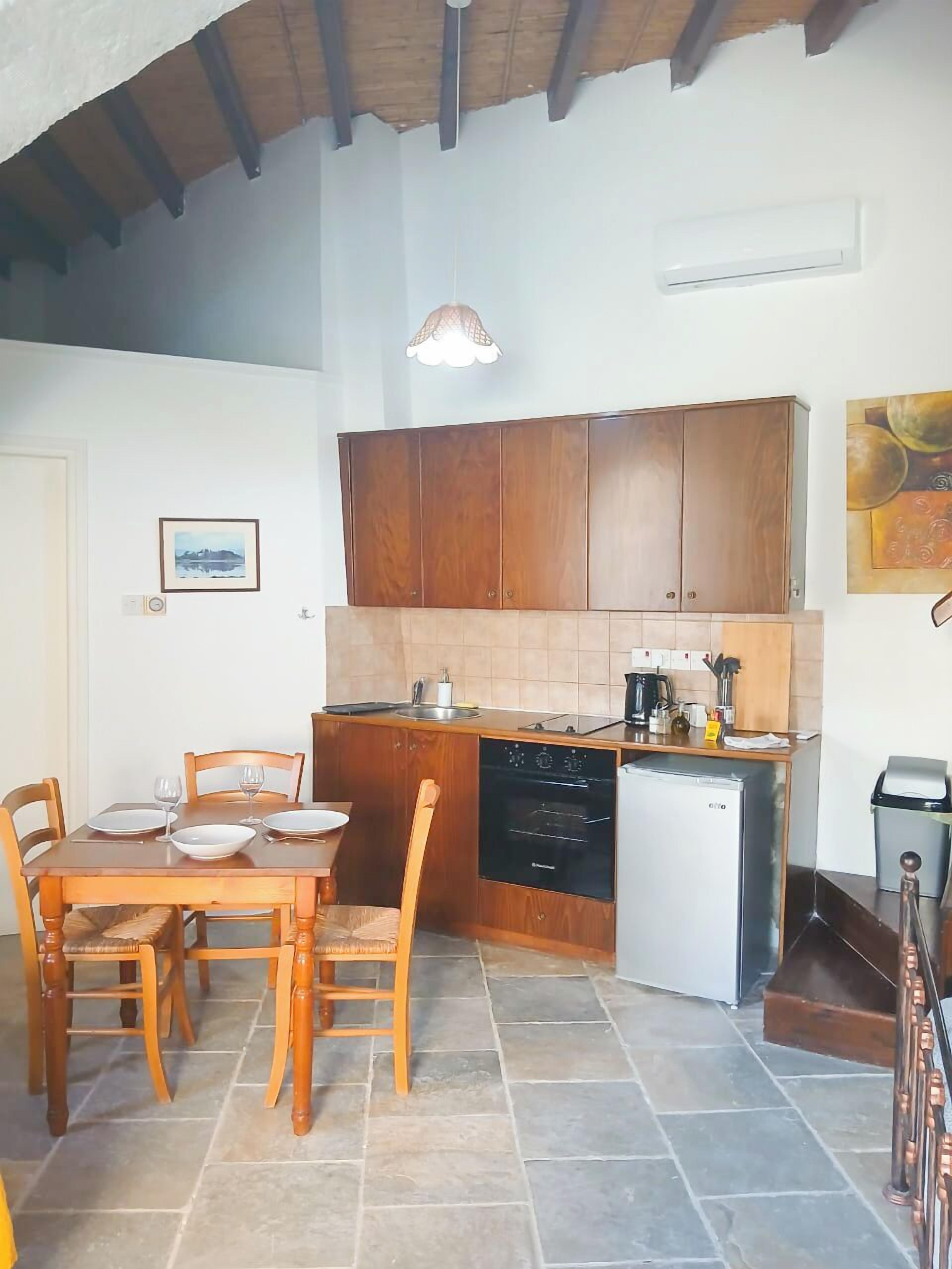 Cocina compacta con armarios de madera, una mesa de comedor con sillas, horno empotrado y un refrigerador pequeño.