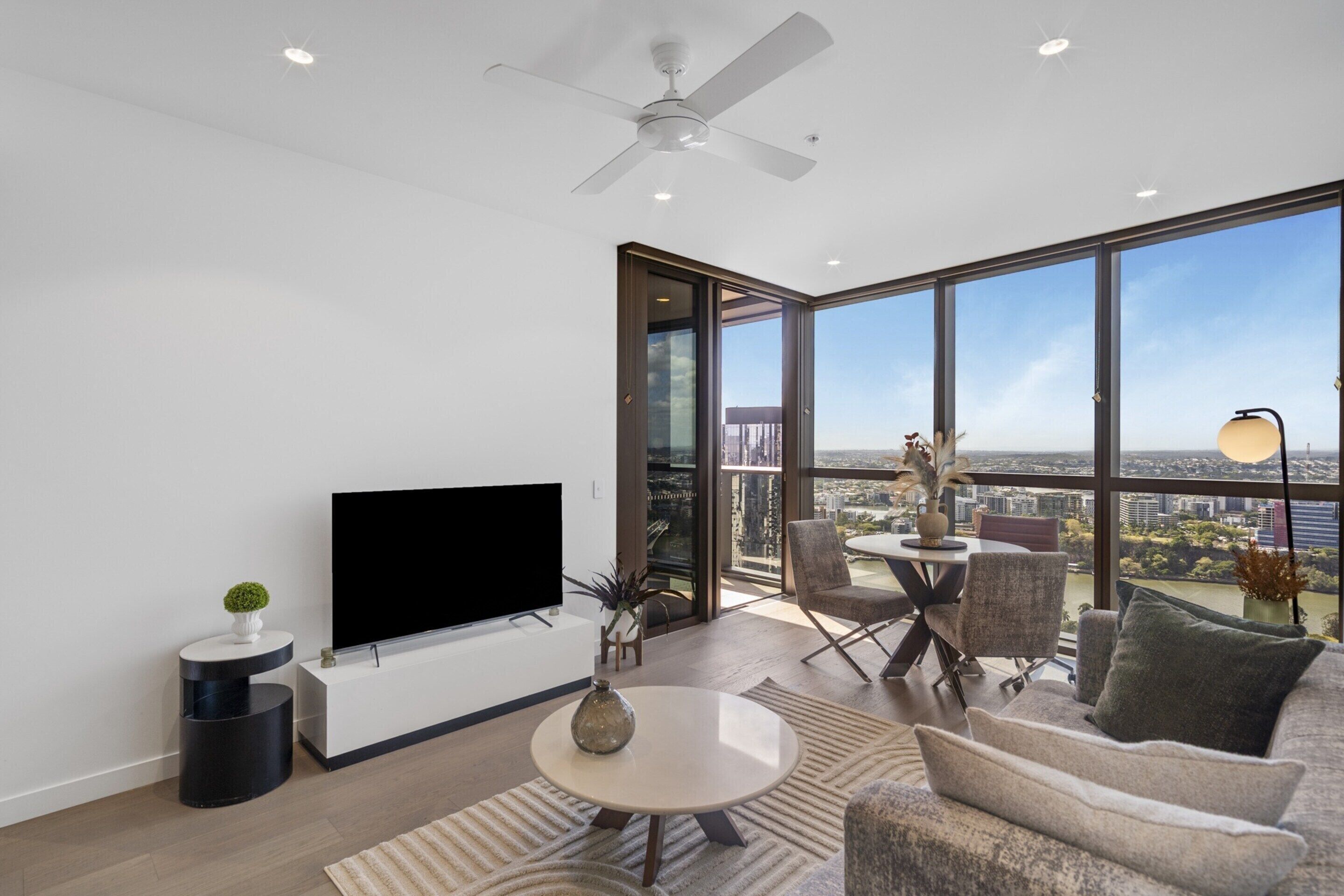 A modern living room with a flat-screen TV, a ceiling fan, a dining area with chairs, and a large window offering a city view.