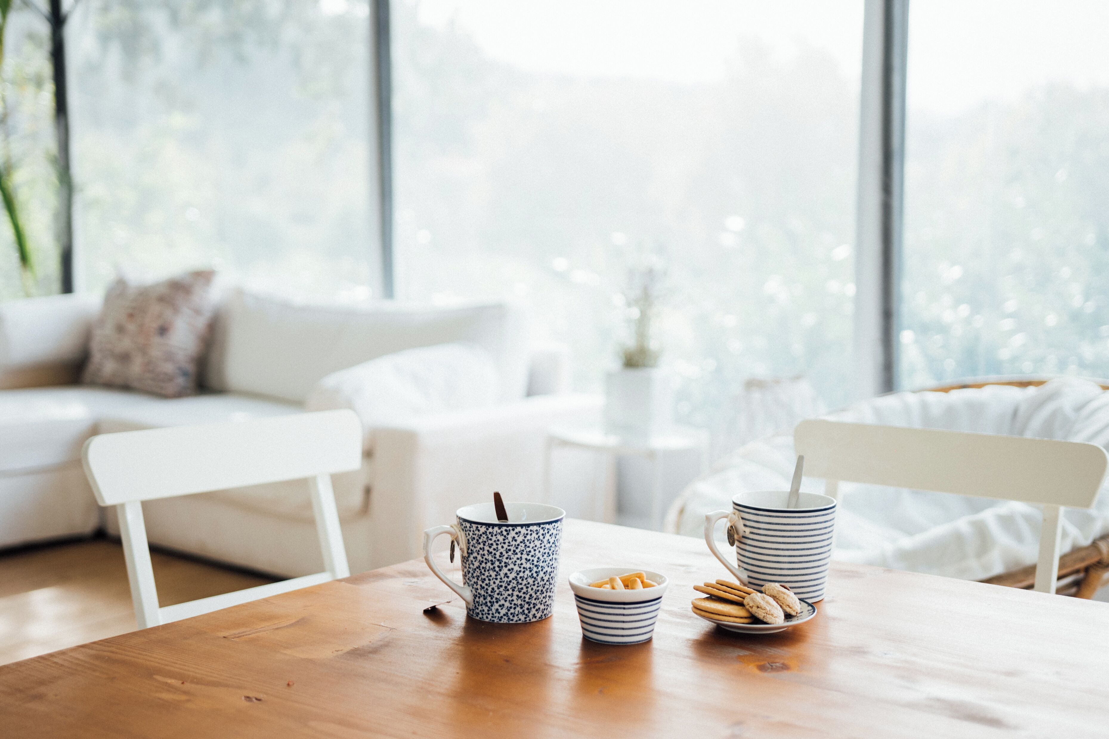 A cozy room with a wooden table, a white chair, a sofa, and a window with a view of trees.