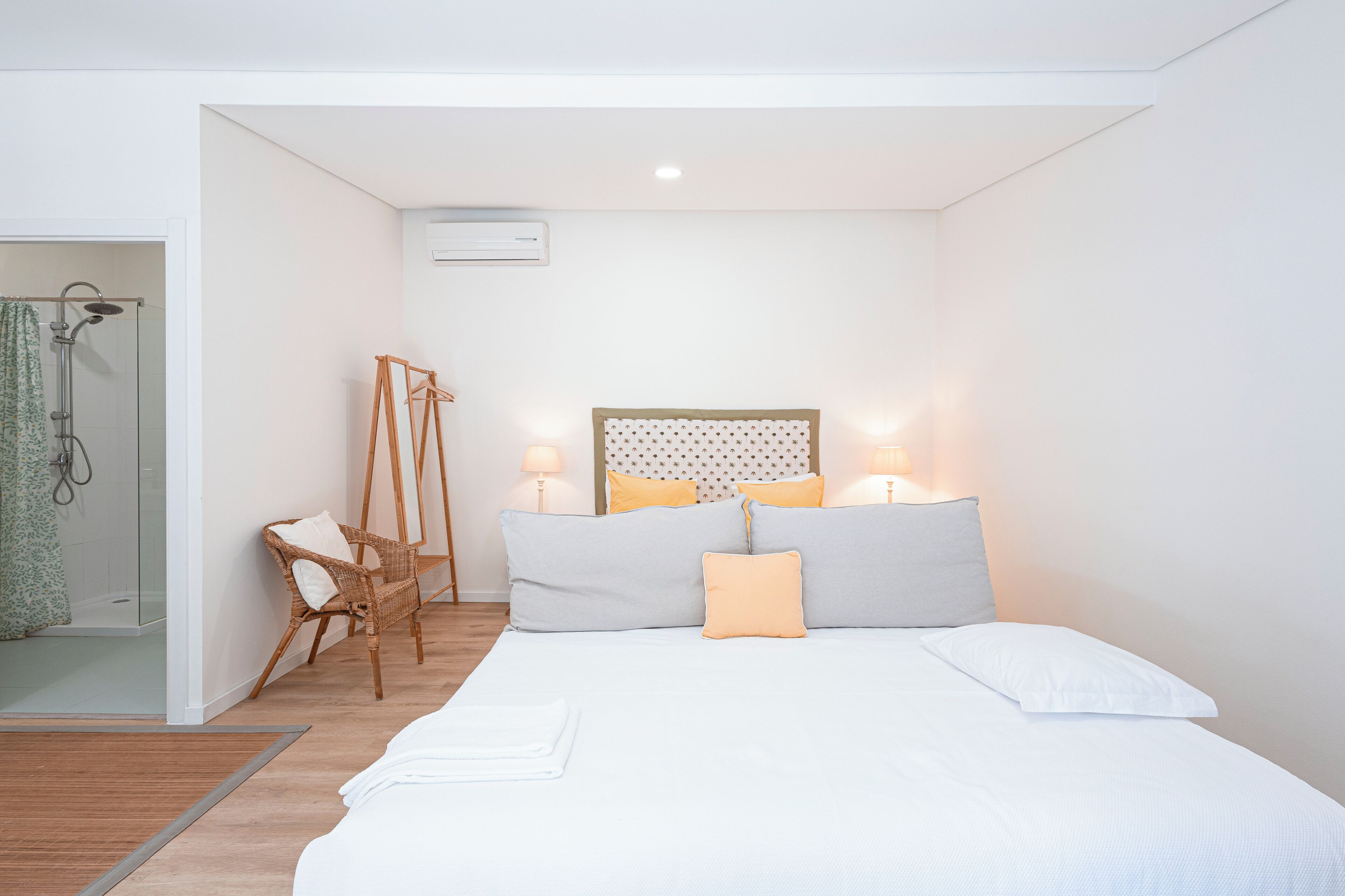 Un dormitorio con cama, lámparas de noche, cabecero, una silla de mimbre y zona de ducha.