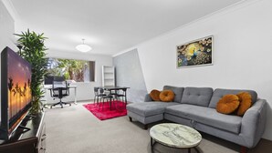 A modern living room with a grey sectional sofa, a round coffee table, and a red rug. There is a painting on the wall and a plant in the corner. - Bright Marrickville Flat With Balcony & Parking (Sydney)