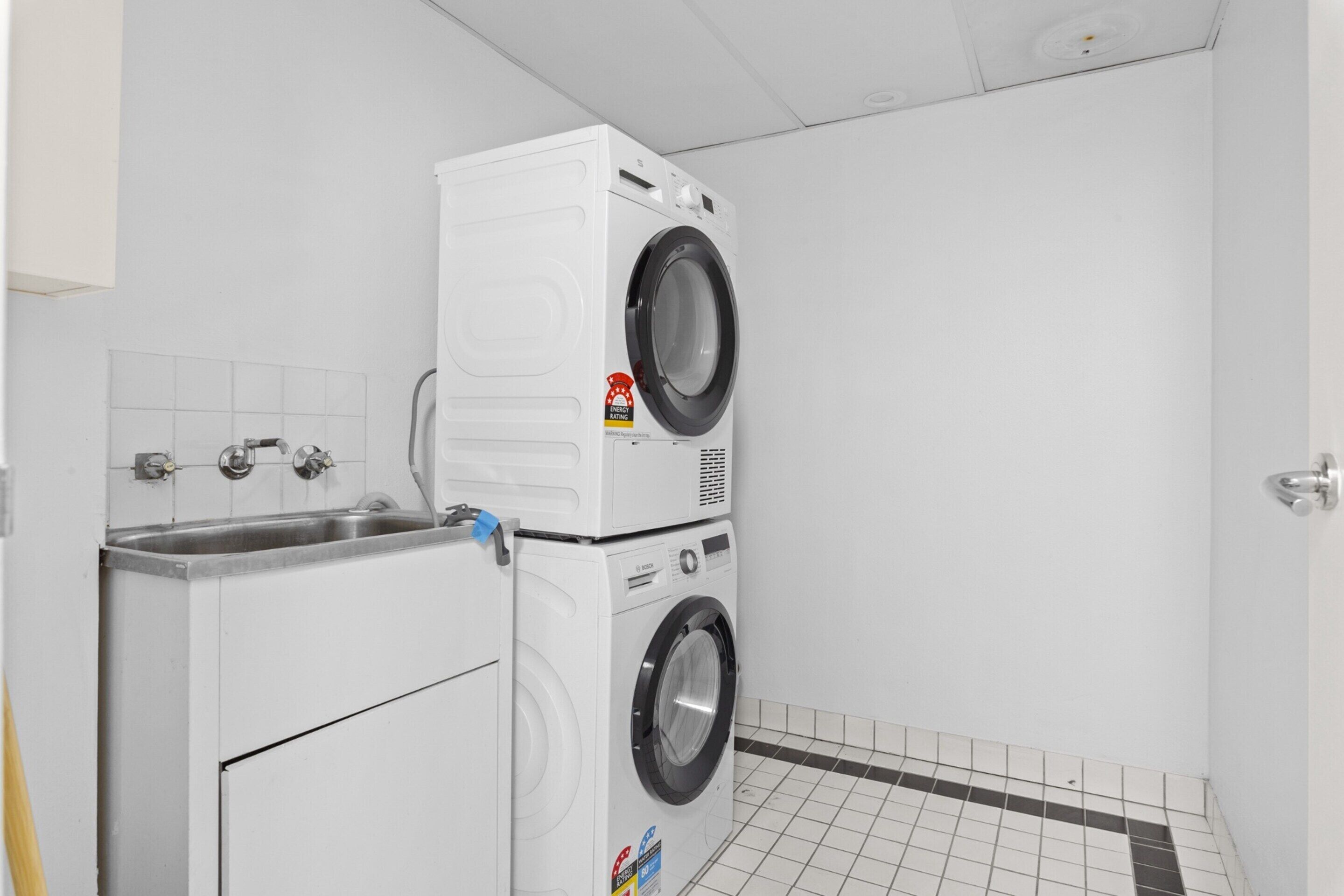Una lavandería con lavadoras apiladas, un lavabo y piso de baldosas.