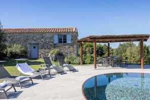 Un bâtiment en pierre avec un toit recouvert de tuiles, une piscine à la bordure d’un bleu éclatant et un espace extérieur aménagé avec des parasols.