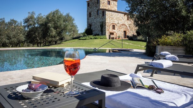 A poolside setting with a stone building in the background, a glass of red wine, a bowl of fruit, and a hat on a table. -  Il Gabbro Luxury by Geco Vacation Rentals (Casole d'Elsa)
