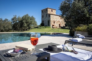 A poolside setting with a stone building in the background, a glass of red wine, a bowl of fruit, and a hat on a table. -  Il Gabbro Luxury by Geco Vacation Rentals (Casole d'Elsa)