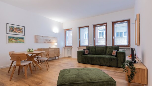 Een moderne woonkamer met een groene bank, een houten eettafel en stoelen. Drie ramen met zonwering, een radiator en een kleine plant op een bijzettafeltje.