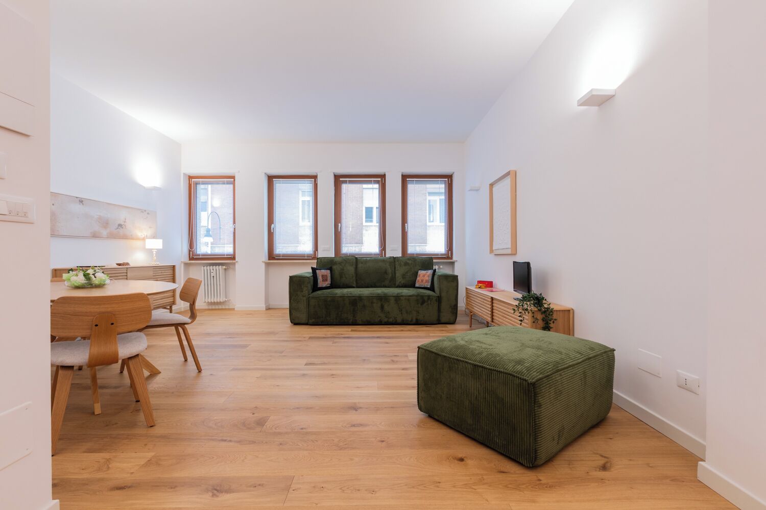 A modern living room with a green sofa, wooden dining table, and chairs. There are four windows providing natural light.