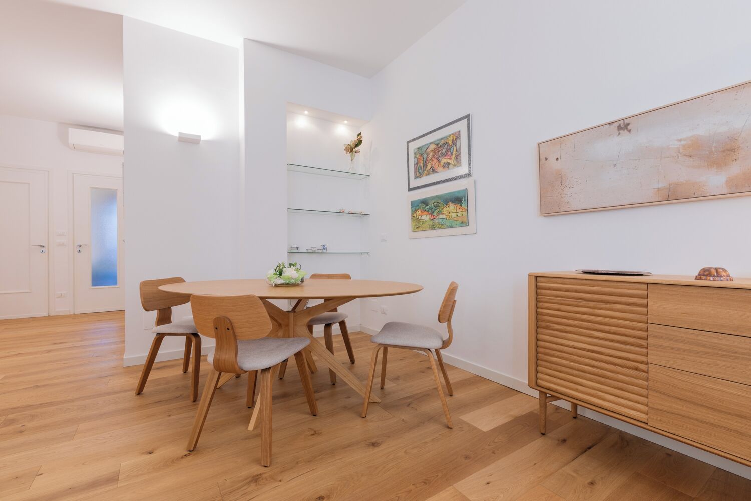 A modern dining area with a wooden table and chairs, a glass shelf with decorative items, and a wooden cabinet with drawers.