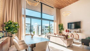 Een moderne woonkamer met een groot raam, een eethoek en een aan de muur bevestigde televisie.