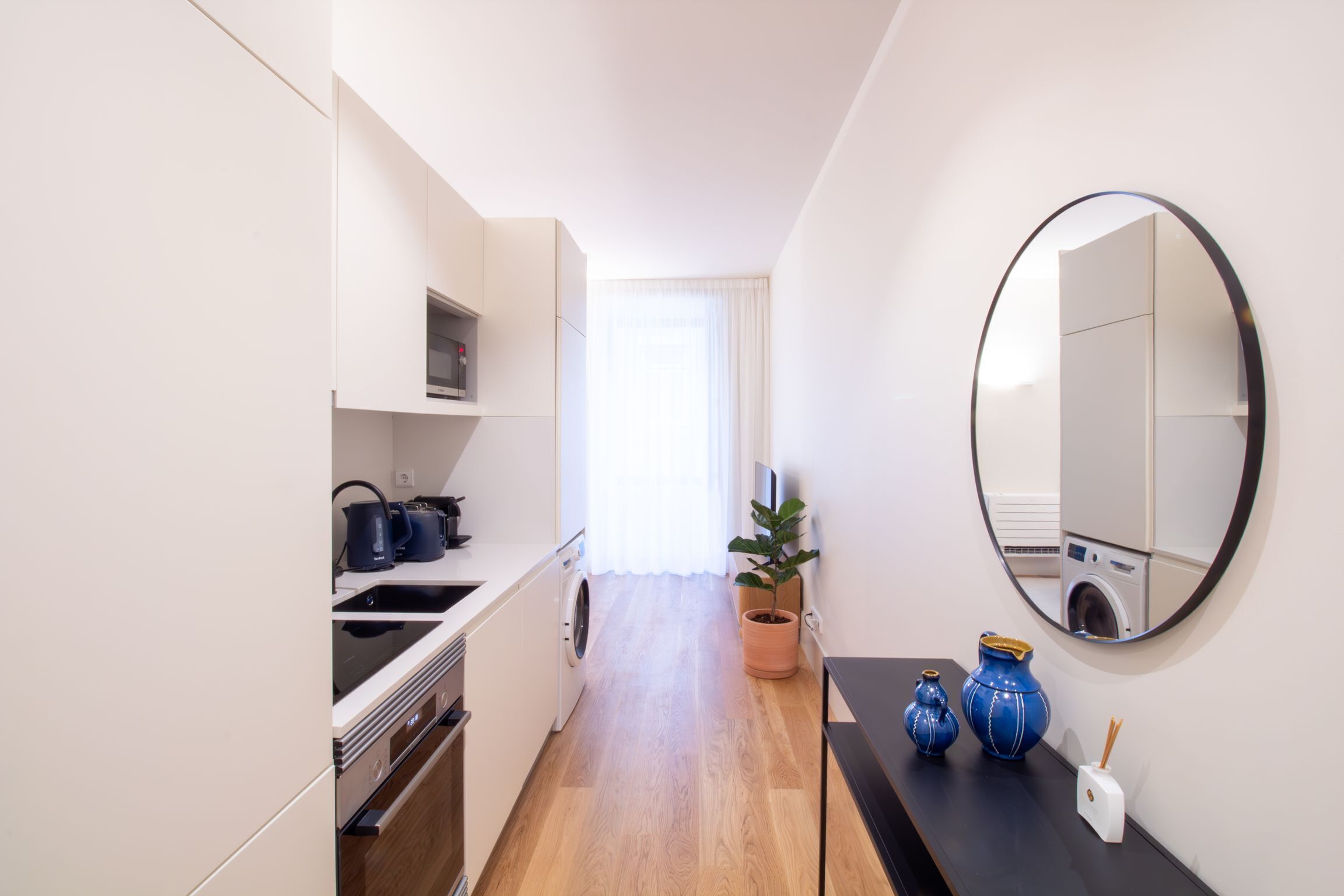 A narrow kitchen with a washing machine, a potted plant, and a mirror on the wall.