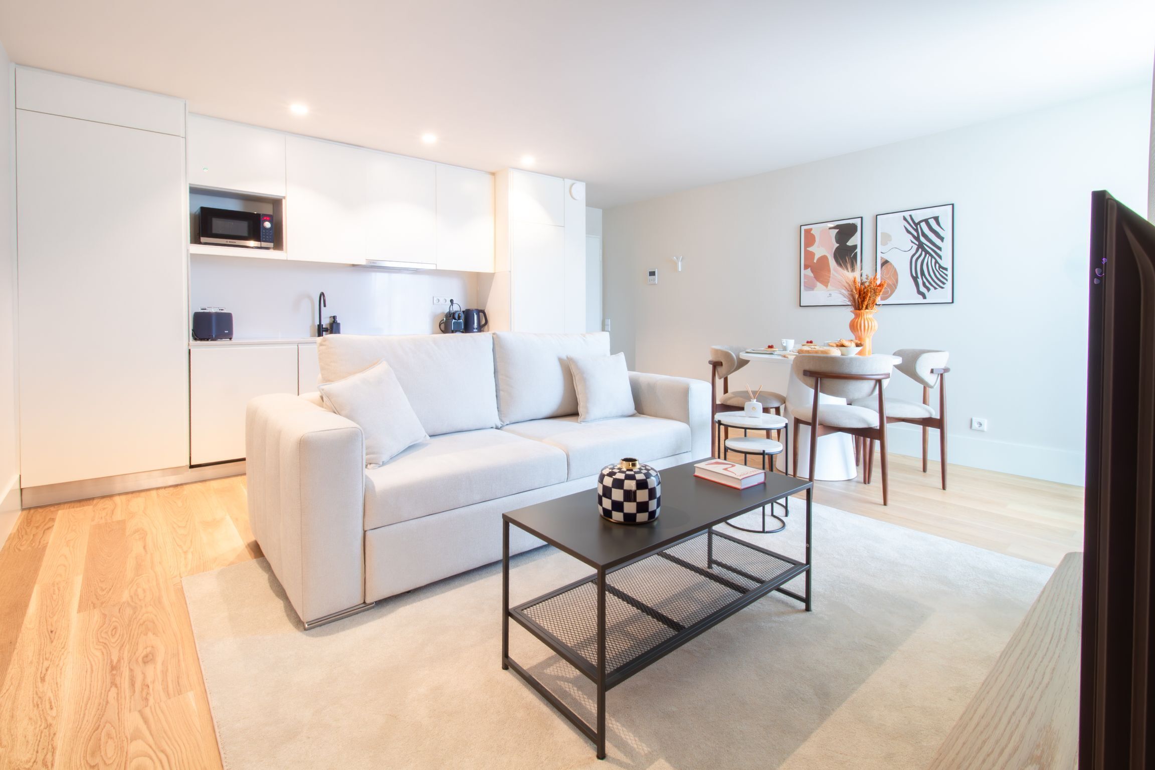 A modern living room with a white sofa, a black coffee table, and a kitchen area with white cabinets and a microwave.
