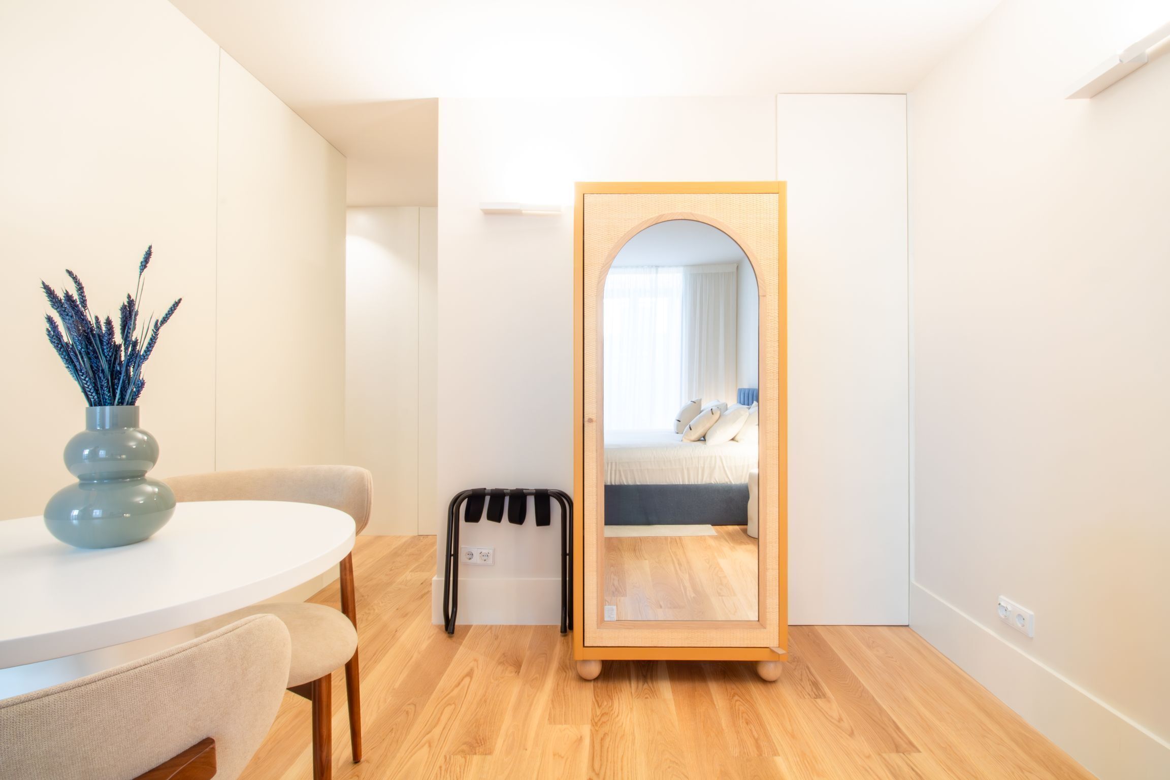 A modern hotel room with a round dining table, a large mirror, a small trash bin, and a bed with a blue headboard.