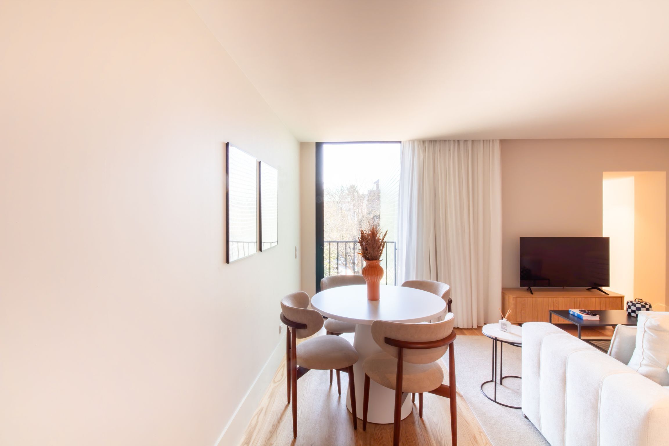 Ein modernes Wohnzimmer mit einem runden Esstisch, weißen Sofas, einem Fernseher und einem großen Fenster mit Vorhängen.