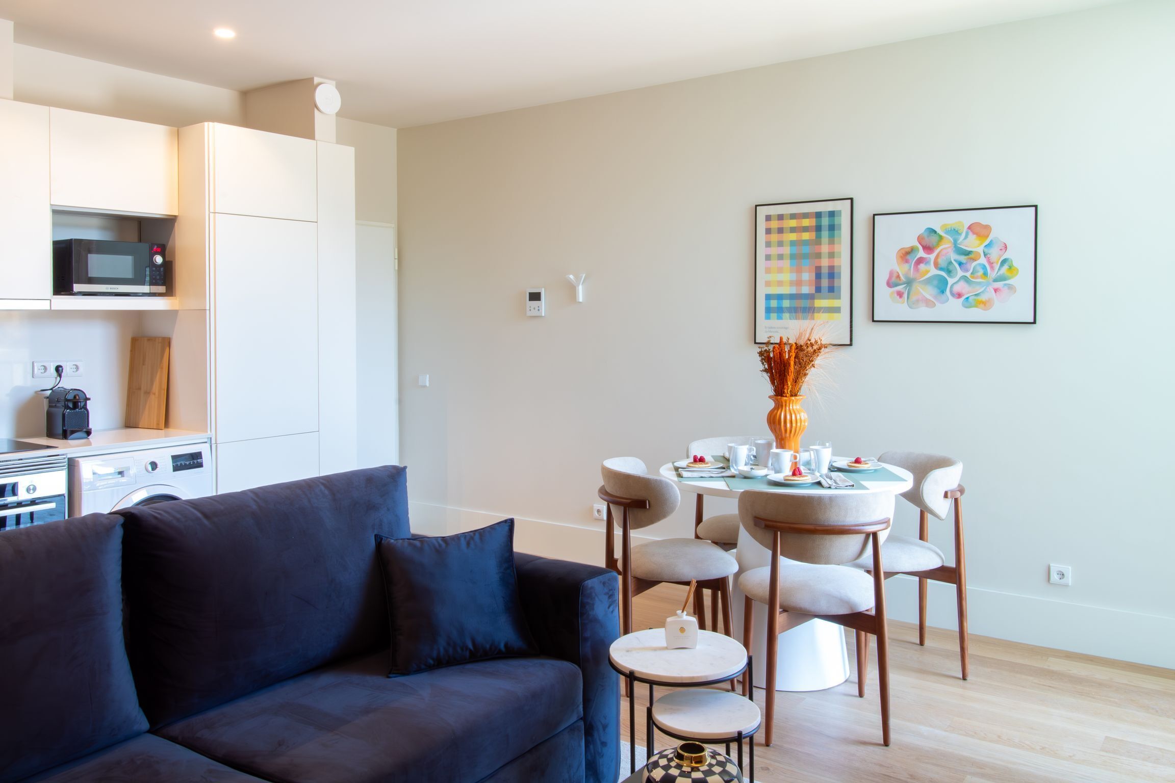 Un salon moderne avec un canapé bleu foncé, un coin repas avec une petite table et des chaises, un four à micro-ondes et un lave-linge.