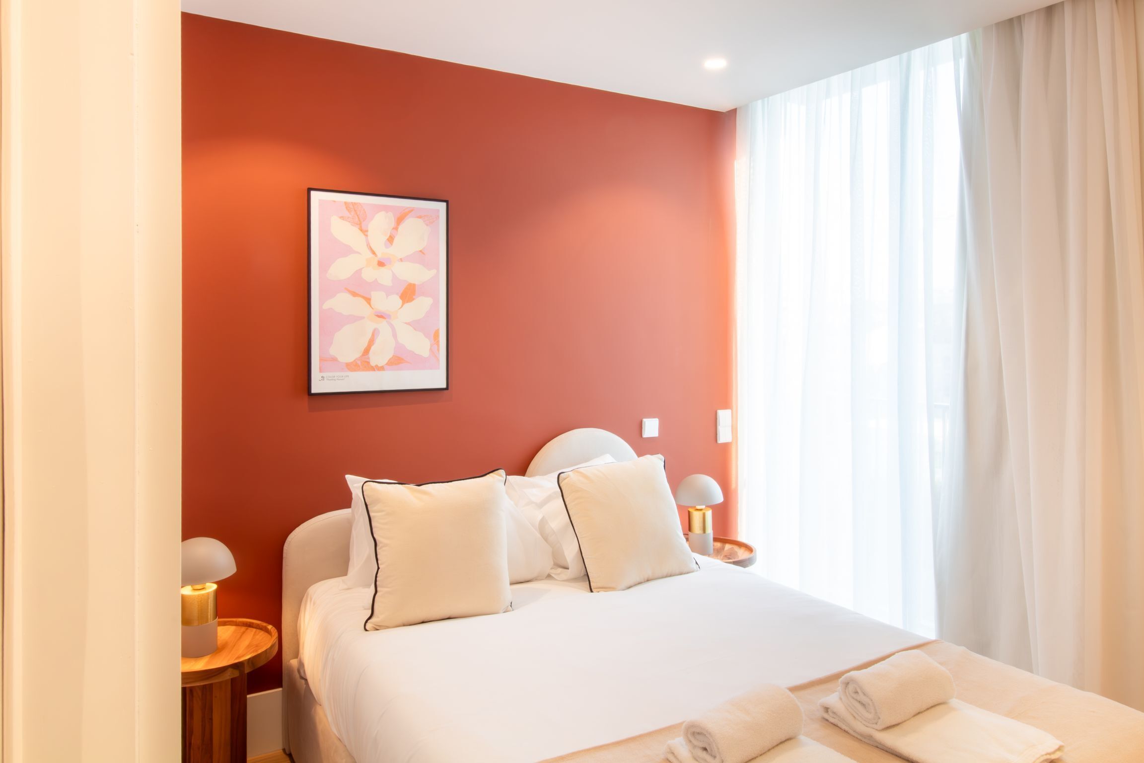 Una habitación de hotel con una cama grande, ropa de cama blanca, un cuadro de flores enmarcado en la pared y una ventana con cortinas de encaje.